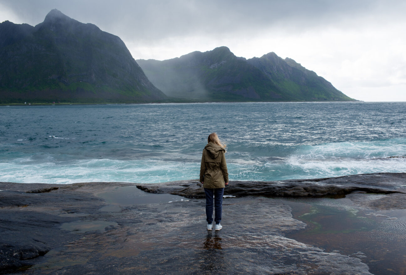 Blond hair girl with a backpack stands on big stones near the water and looks at the ocean. Waves, splashing. Enjoy the moment, relaxation. Wanderlust. Travel, adventure, lifestyle. Explore Norway Safe Places to Travel Alone As a Woman: Empower Your Solo Adventures